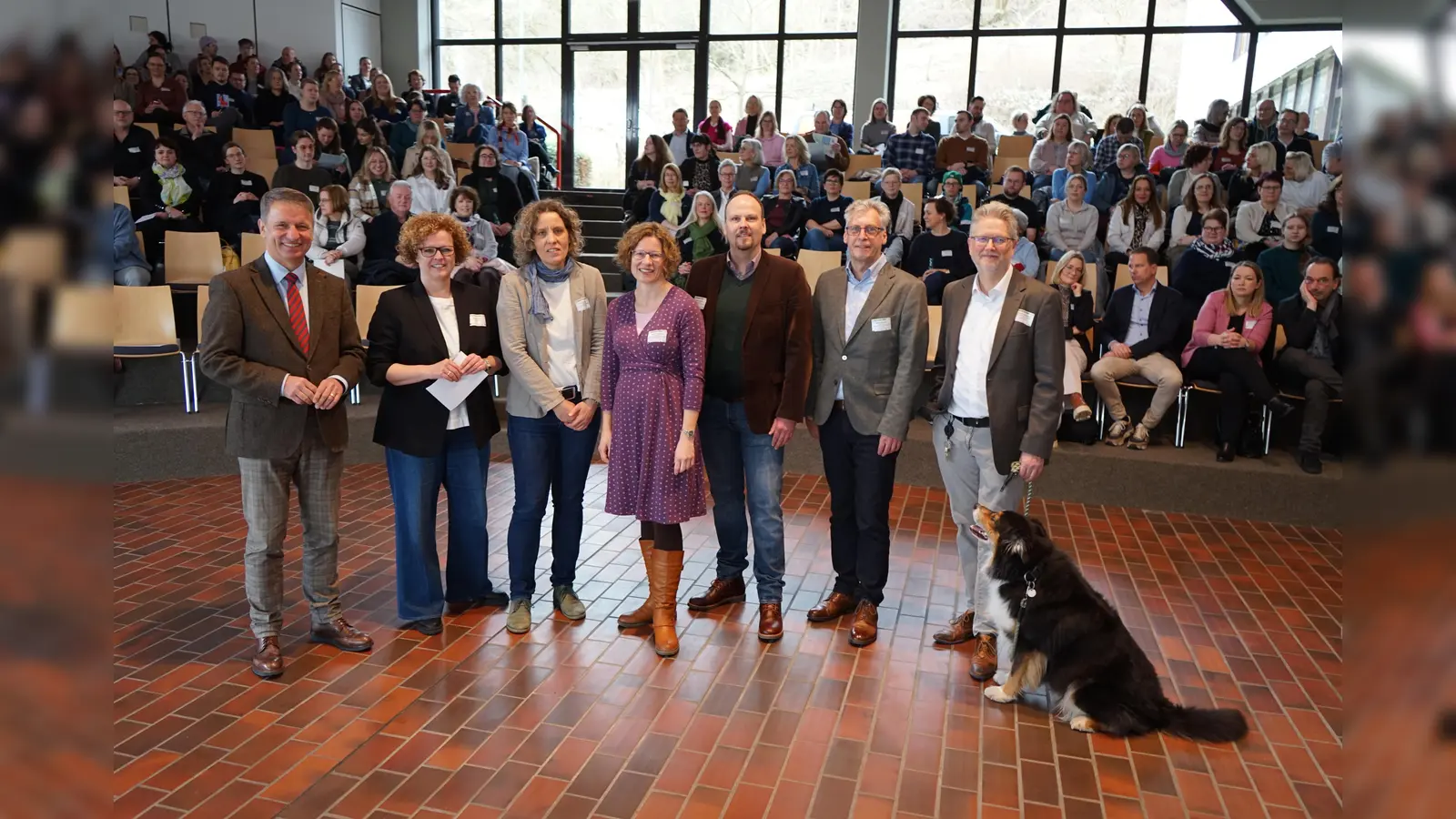 Freude über rund 250 Teilnehmende am Fachtag „Junge Menschen mit psychischen Belastungen in der beruflichen Orientierung“ in Höxter: (v.l.) Landrat Michael Stickeln, Moderatorin Susanne Stork, Caroline Rieger, Katrin Niemann (beide Kommunalen Koordinierungsstelle Übergang Schule-Beruf des Kreises Höxter), Dominic Gehle (Leiter der Abteilung Bildung und Integration des Kreises Höxter), Stefan Fellmann (Dezernent für Schulen, Ordnung, Soziales und Recht der Stadt Höxter) und Referent Dr. Dirk Dammann (Leiter der Klinik Albert-Schweizer-Therapeutikum in Holzminden) mit Hund Bagira. (Foto: Kreis Höxter)